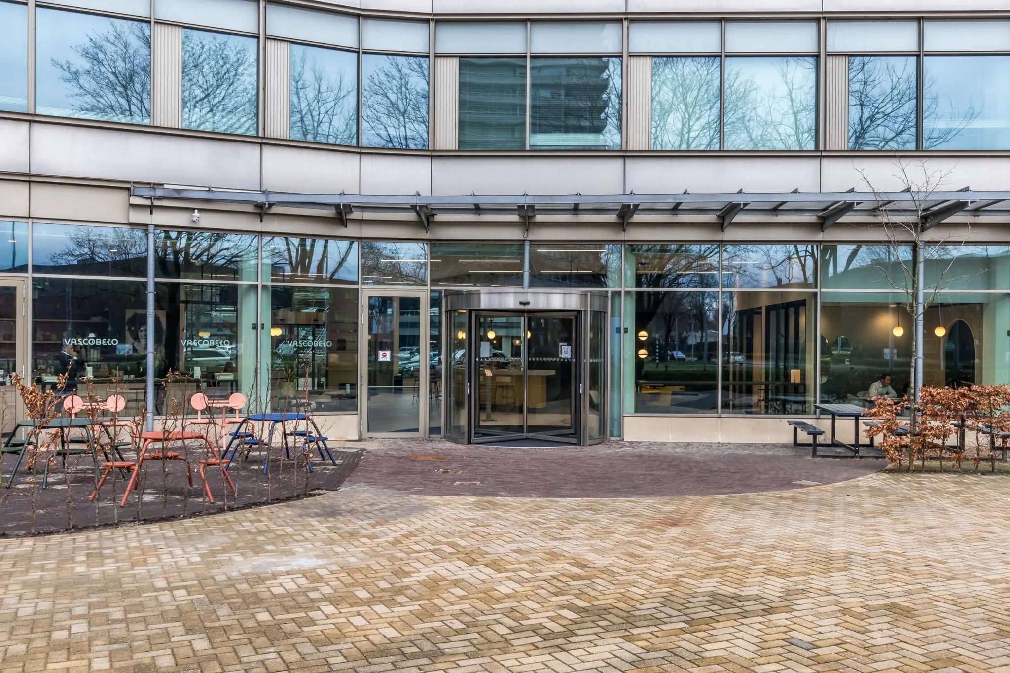 Vooraanzicht van een modern kantoorgebouw aan de Vliegend Hertlaan met glazen gevel, entree en buitenterras met stoelen en tafels.